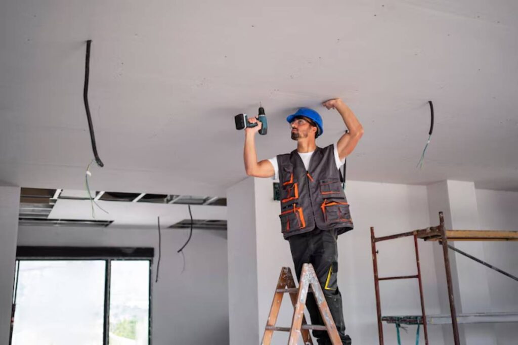 A worker stands on a ladder, using a power drill to install ceiling fixtures in a room with exposed wiring and a scaffold.