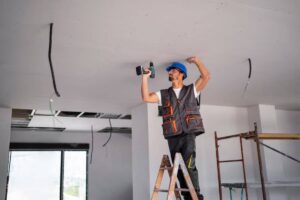 A worker stands on a ladder, using a power drill to install ceiling fixtures in a room with exposed wiring and a scaffold.