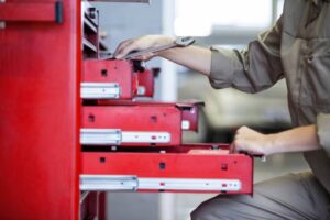 A mechanic in a beige jumpsuit organizes tools in a red toolbox, holding a wrench and accessing drawer compartments.