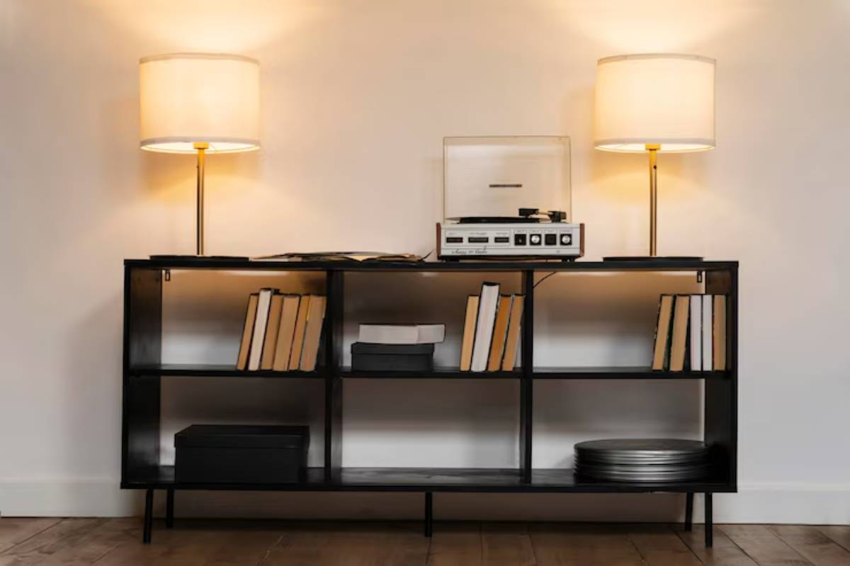 A modern black shelf holds books and a vintage record player, flanked by two soft-glow lamps against a white wall.