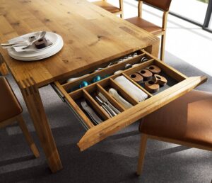 A wooden dining table with a drawer open, showcasing neatly organized cutlery, plates, and various wooden utensils, with chairs nearby.