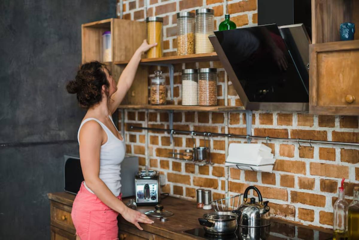 A woman in a tank top reaches for a jar on a wooden shelf in a rustic kitchen with exposed brick walls and a cluttered countertop.