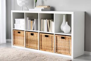 A modern white bookshelf with six compartments, showcasing books and decorative vases, alongside woven storage baskets.