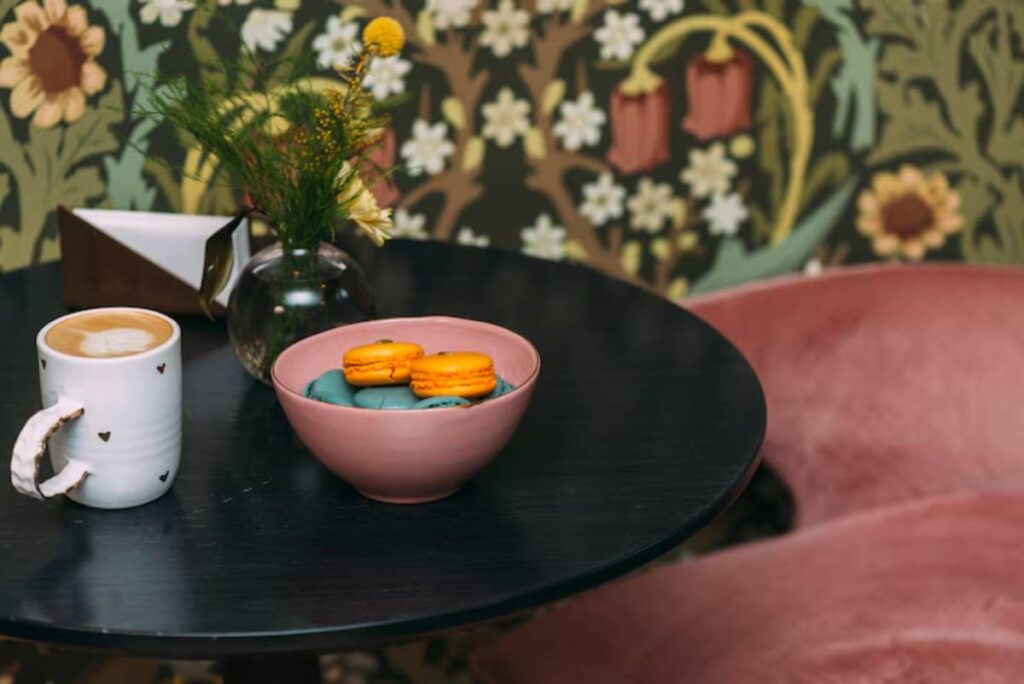 A cozy table setting features a white mug of coffee, a pink bowl of colorful macarons, and a vase of flowers against a floral backdrop.