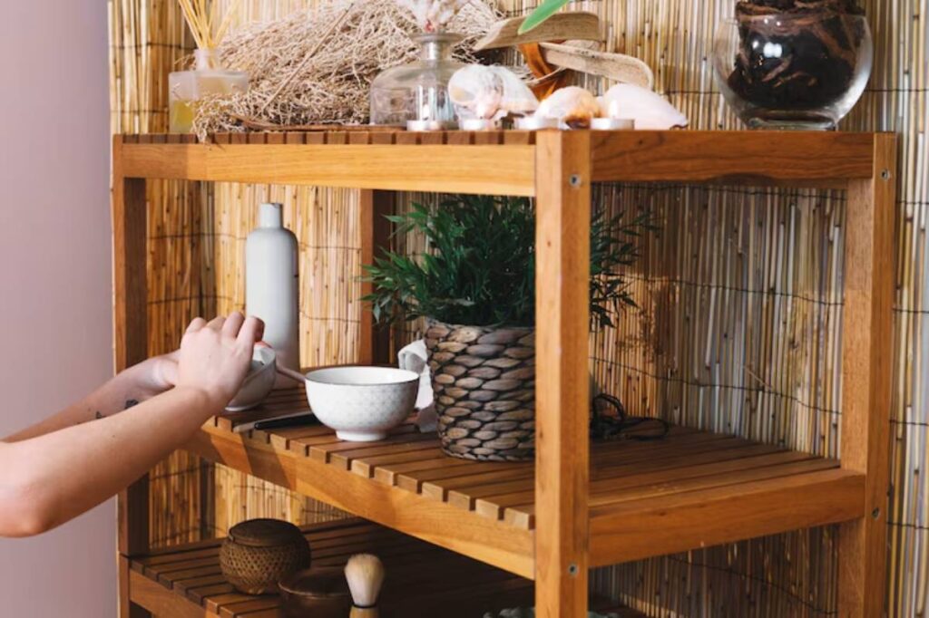 A person arranging decorative items on a wooden shelf with plants, bowls, and various accessories against a bamboo backdrop.