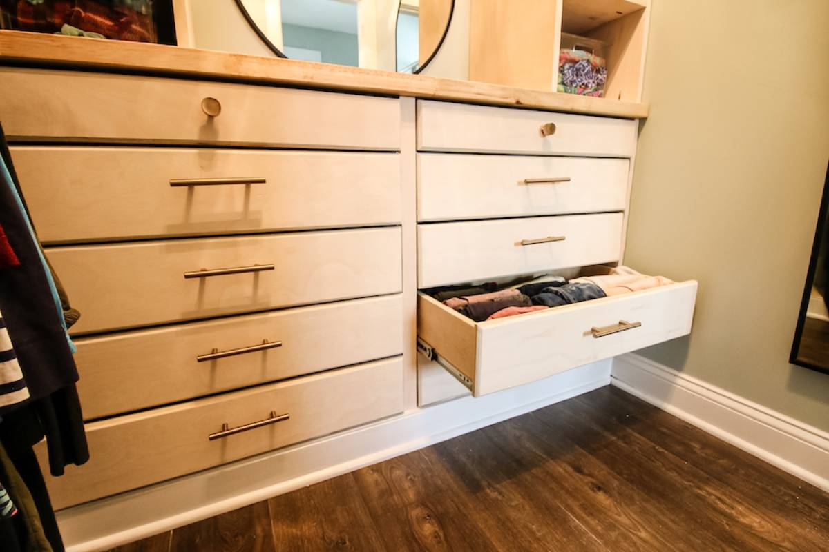 A modern dresser with multiple drawers, one open showcasing neatly folded clothes, beside a mirror and colorful clothing hanging nearby.