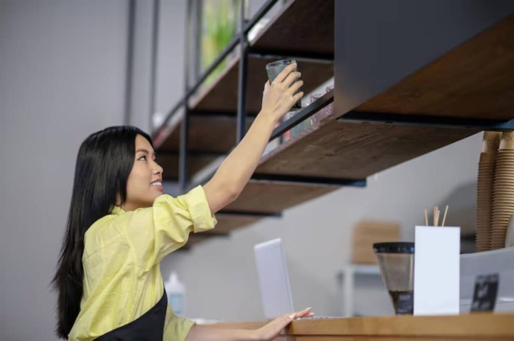 A woman in a yellow shirt reaches for a glass on a wooden shelf above a counter, surrounded by other café items.
