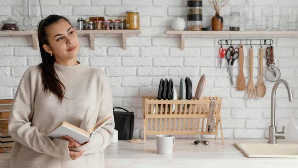A person stands in a modern kitchen holding an open book, surrounded by white brick walls, shelves of jars, and kitchen utensils.