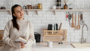A person stands in a modern kitchen holding an open book, surrounded by white brick walls, shelves of jars, and kitchen utensils.