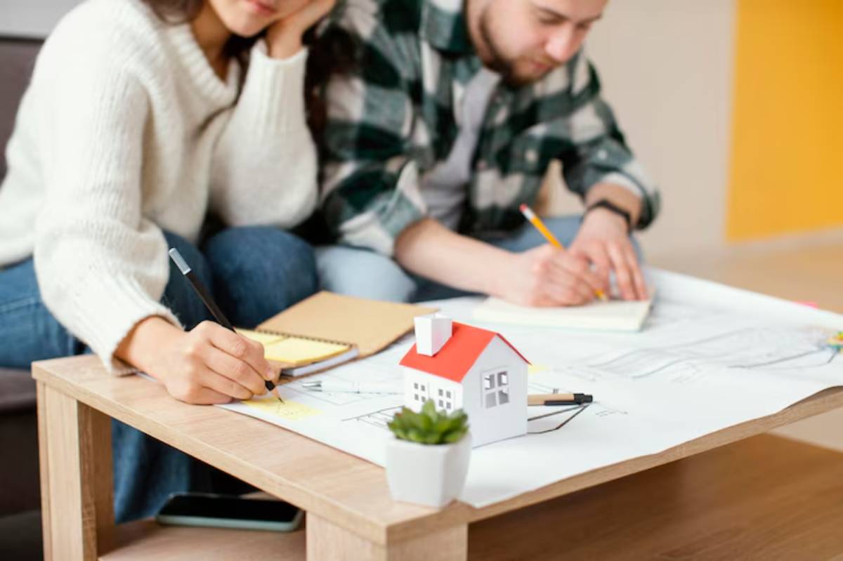 A tabletop scene featuring two individuals sketching architectural plans, with a small model house and notebook among their materials.