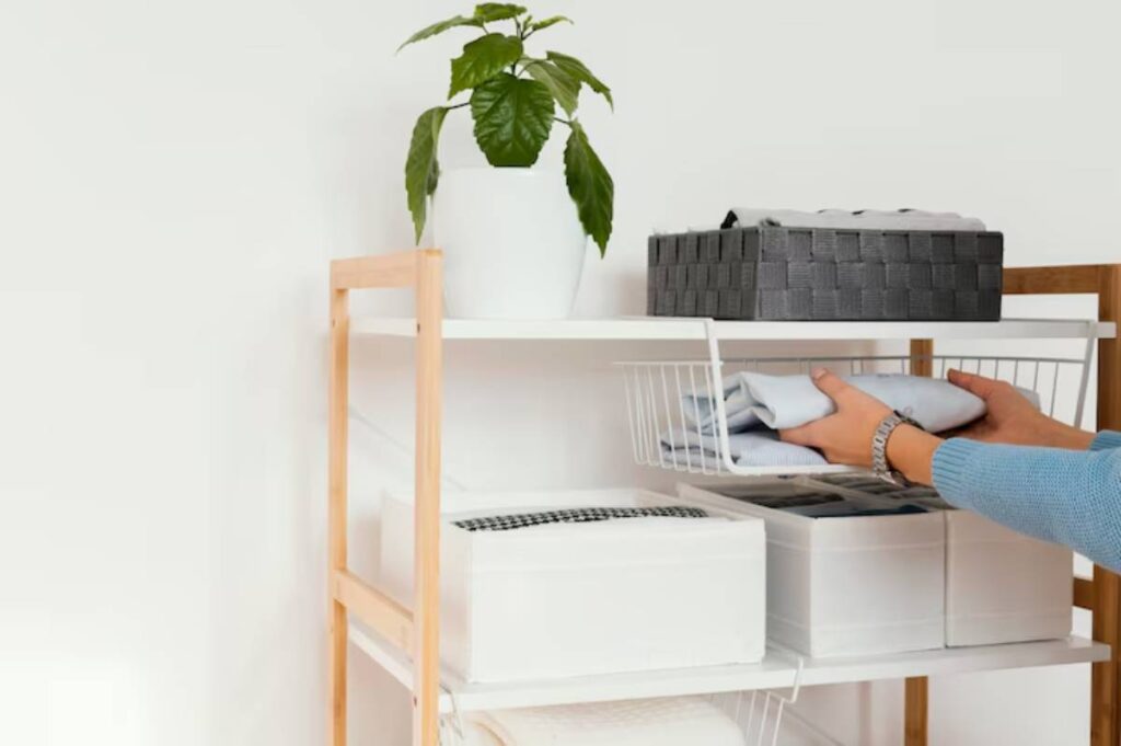 A person organizes folded clothes in a white wire basket on a wooden shelf, accompanied by a potted plant and decorative storage boxes.