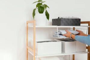 A person organizes folded clothes in a white wire basket on a wooden shelf, accompanied by a potted plant and decorative storage boxes.