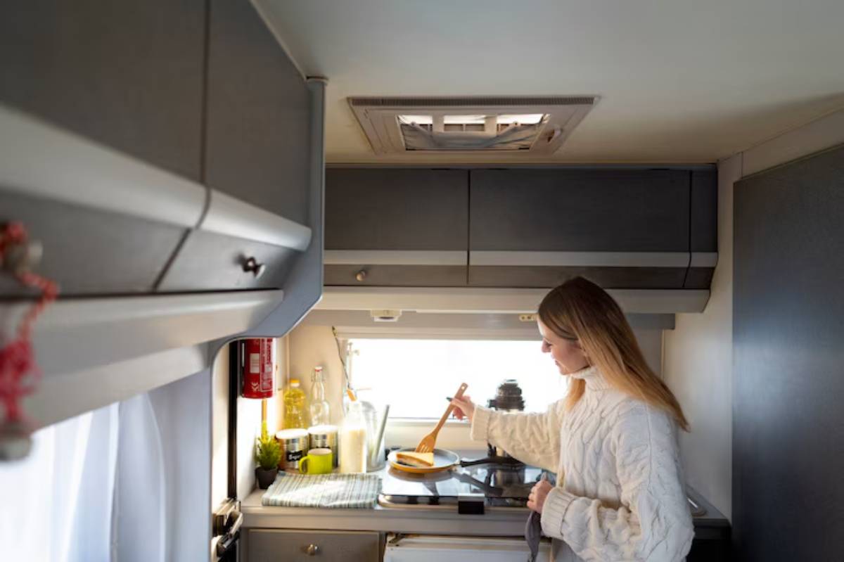 A woman stirs food in a modern camper kitchen, featuring gray cabinets and a bright window with natural light.