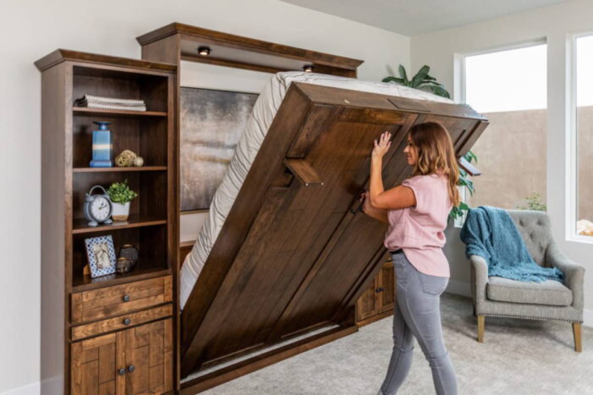 A modern dormitory with two sets of bunk beds in warm wood tones. One occupant relaxes on a lower bunk while another climbs a ladder.