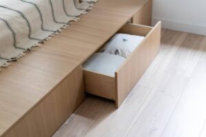A modern wooden bed with a light blanket draped over it, revealing a drawer underneath containing a patterned pillow.
