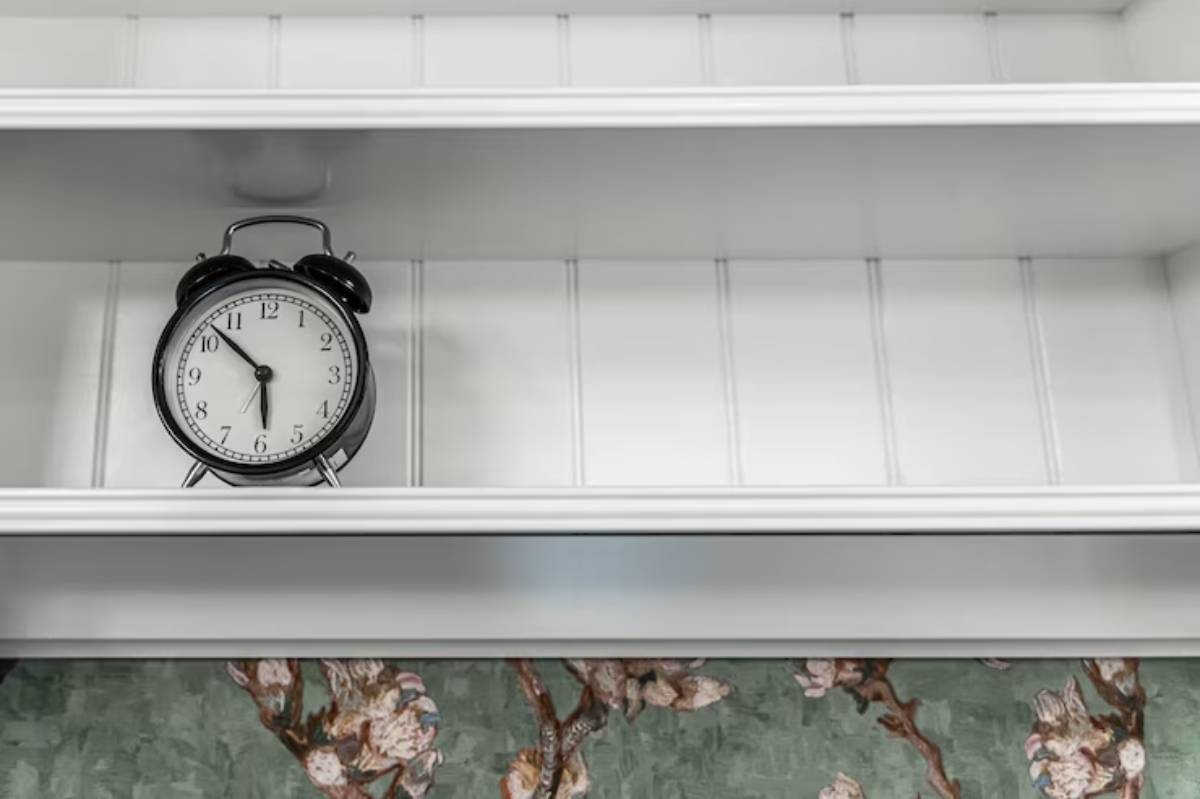  A black alarm clock sits on a white shelf, above floral-patterned wallpaper, displaying the time at 10:10.