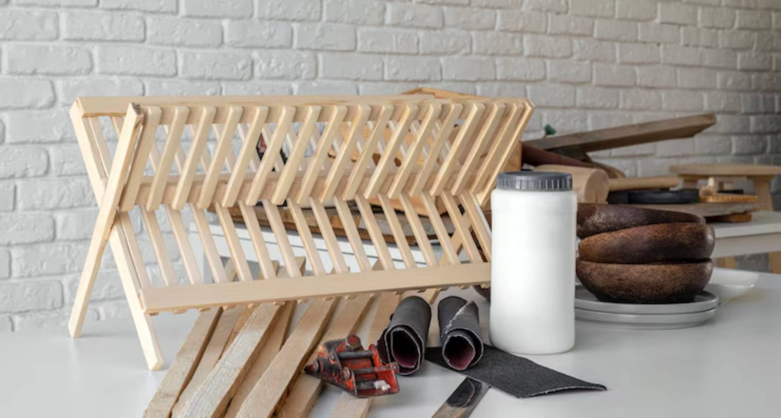A wooden drying rack with various woodworking tools, a white container, and unfinished wood pieces on a light surface against a brick wall.