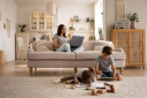 A cozy living room with a couch and large rug, where a woman works on a laptop while children draw and play on the floor.