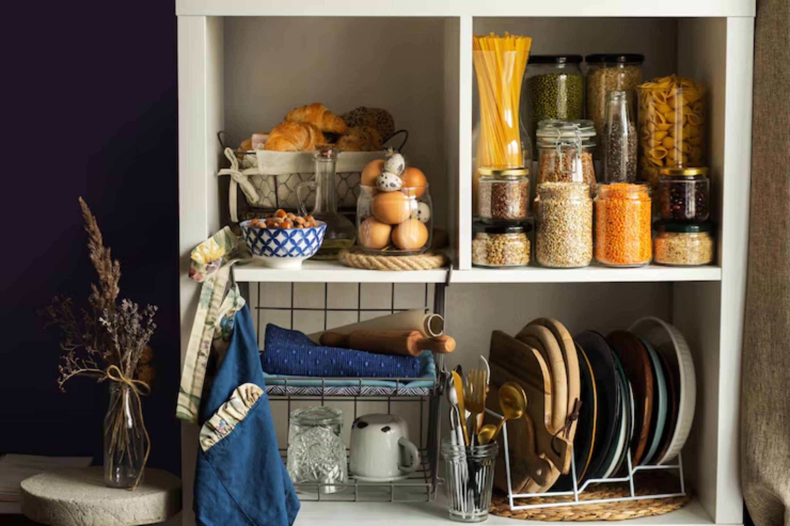 A stylish, organized kitchen shelf displaying jars of grains, pasta, eggs, croissants, and utensils, with decorative touches.