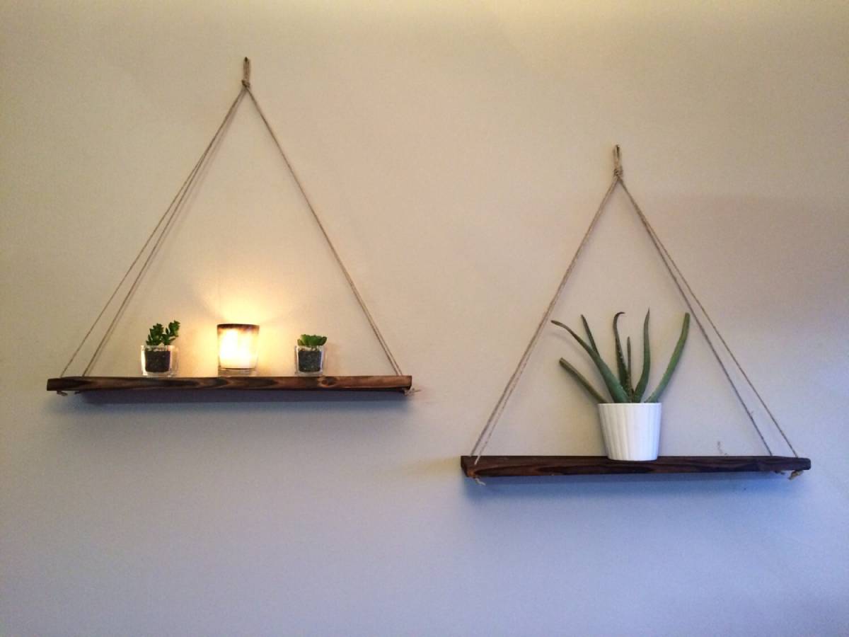 Two triangular wooden shelves are hung on a wall, featuring small plants and a glowing candle on the left shelf, and an aloe plant on the right.