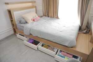 Cozy bedroom featuring a bed with storage drawers underneath, light-colored bedding, a plush toy, and sunlight filtering through curtains.