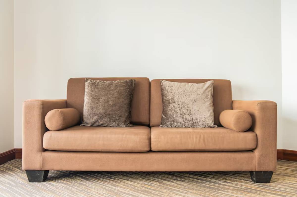 A cozy brown sofa with two square cushions and a round bolster cushion, set against a plain wall and textured carpet. 