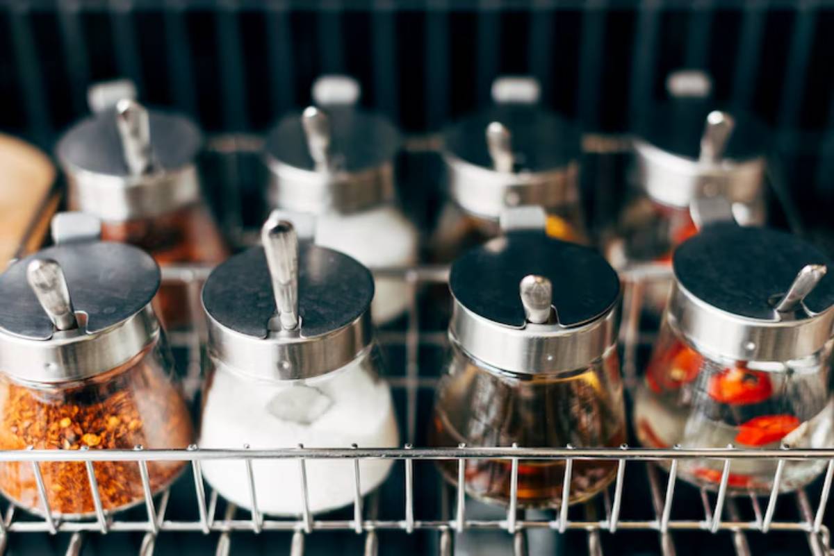  A wire rack holds various glass spice jars with metal lids, featuring ingredients like chili flakes, salt, and infused oils.