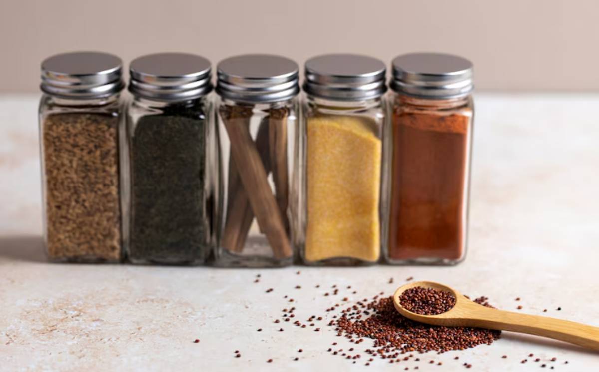  Five glass spice jars with silver lids line up, featuring various spices and a wooden spoon scooping vibrant seeds on the surface.
