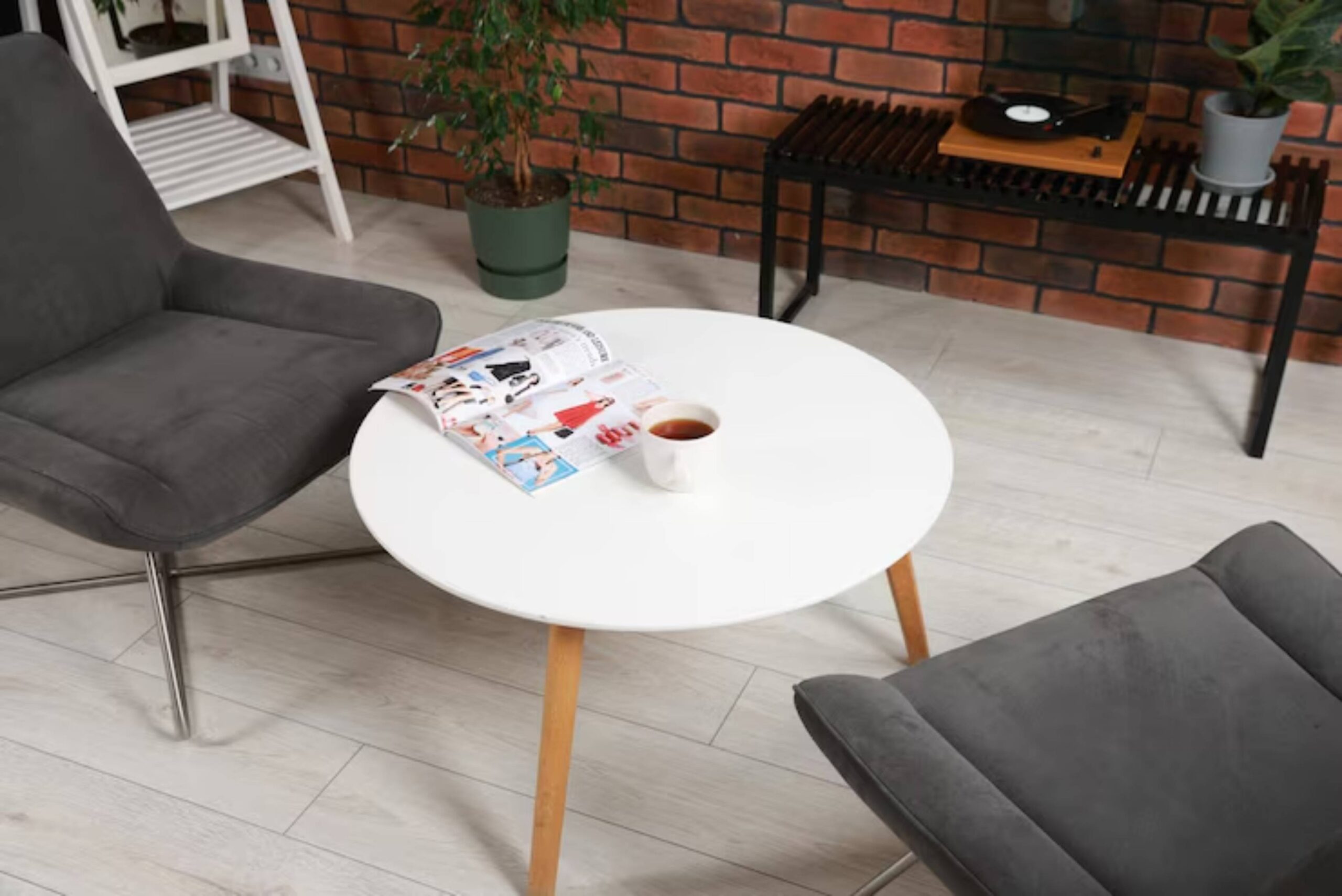 A cozy seating area with a gray chair, round white table, a cup of tea, and a magazine, set against a brick wall.