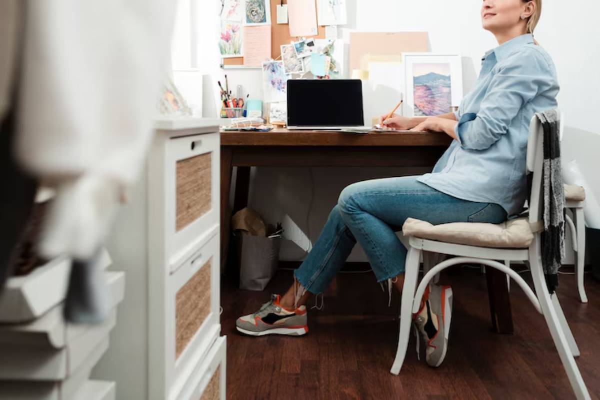 A person sits at a wooden desk with a laptop, writing notes, surrounded by creative supplies and a cozy home office environment.