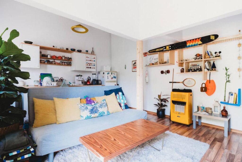 Cozy living room with a blue sofa, colorful cushions, a wooden coffee table, and decorative shelves, featuring plants and vintage items.