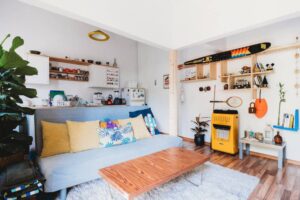 Cozy living room with a blue sofa, colorful cushions, a wooden coffee table, and decorative shelves, featuring plants and vintage items.