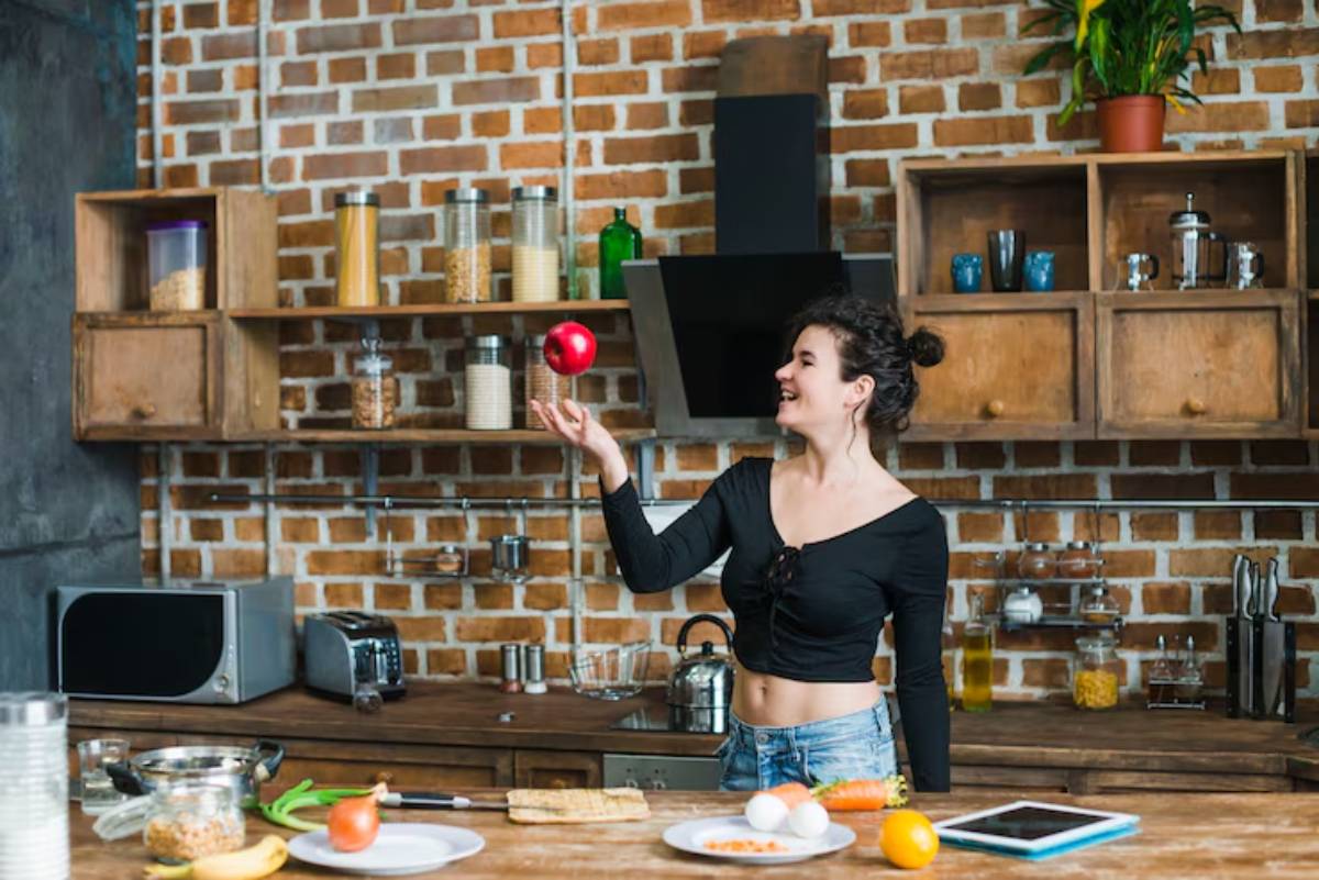 A woman in a black top juggles a red apple in a rustic kitchen, surrounded by fresh vegetables and cookware on a wooden table. 